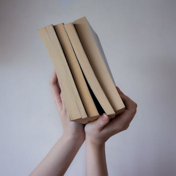 A close-up image of hands holding a stack of paperback books against a neutral backdrop.